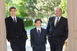 two boys with their father in formal clothes leaning in doorway with greenery in the background